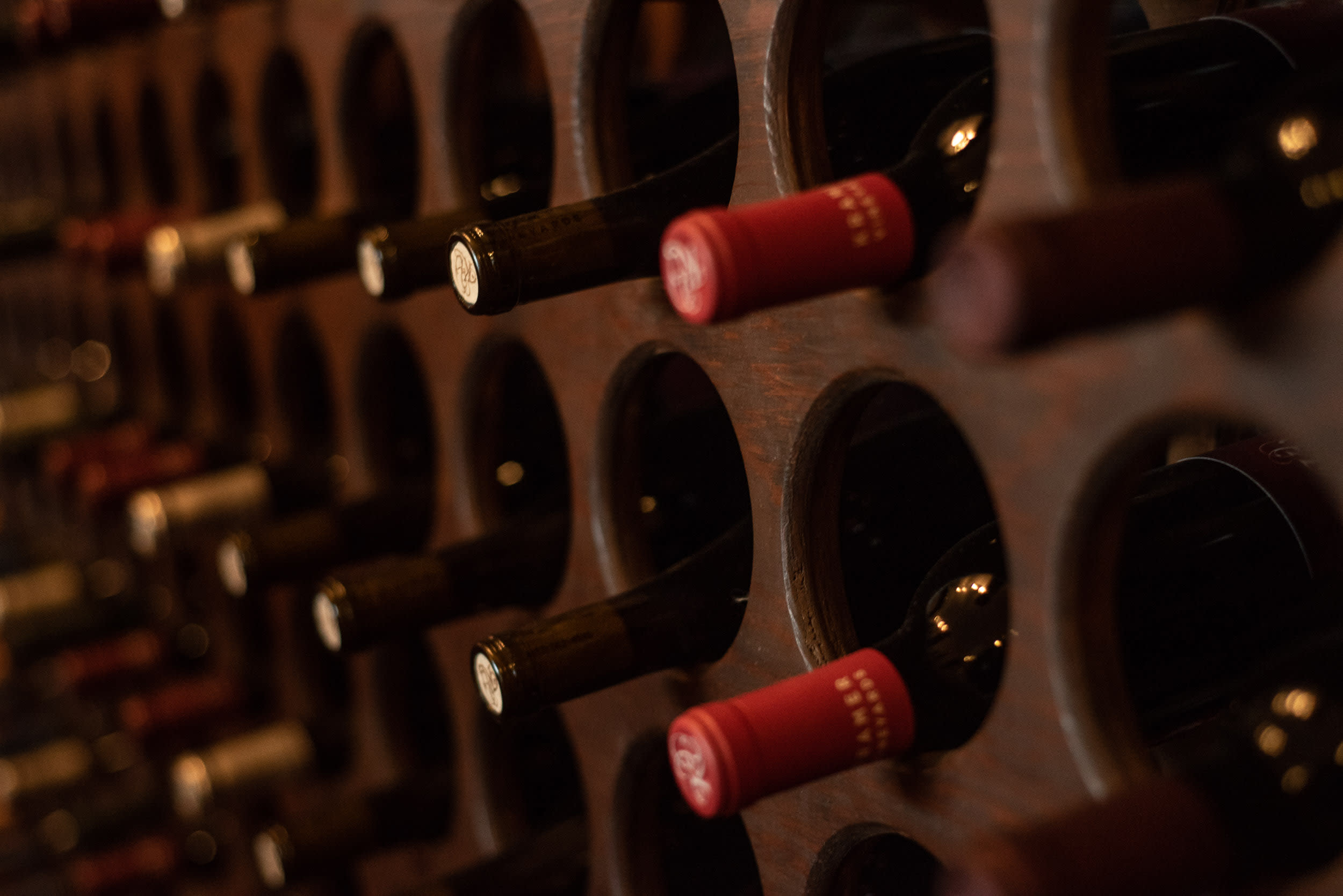 Close-up of wine bottles stored horizontally in a dark wooden wine rack, with visible red and black caps.